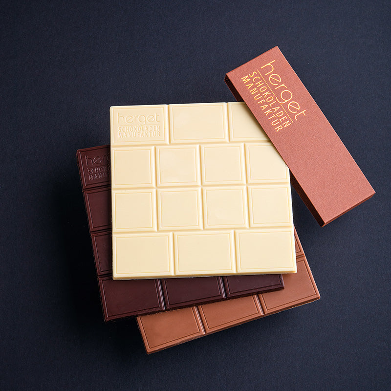 Produktabbildung von drei Schokoaldentafeln in weiß Vollmilch und Zartbitter gestapelt. außerdem eine braune Papierbanderole mit Herget Schokoladen Logo